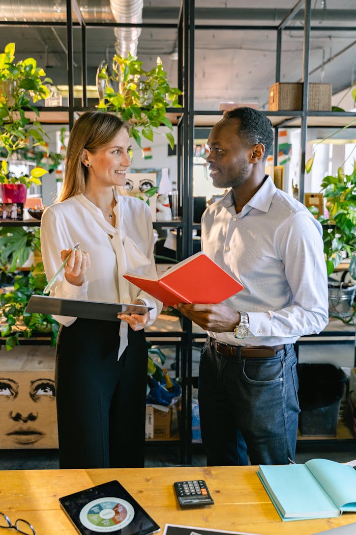 Two colleagues discussing work in a modern office with plants, using notebooks and clipboards.
